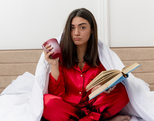 毯子穿着红色睡衣的年轻美女坐在床上 裹着毯子 端着一杯咖啡和一本书 看着灯光背景下卧室里的摄像机 很不高兴室内坐着包