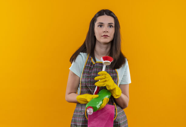 女孩穿着围裙 戴着橡胶手套 拿着刷子和清洁用品的年轻漂亮女孩严肃地看着一旁橙色手套靠边