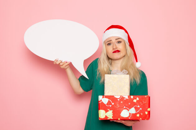 年轻的女性正面图年轻女性手持圣诞礼物和白色标志在粉色墙上的女性礼物雪地彩照新年假期女人雪微笑