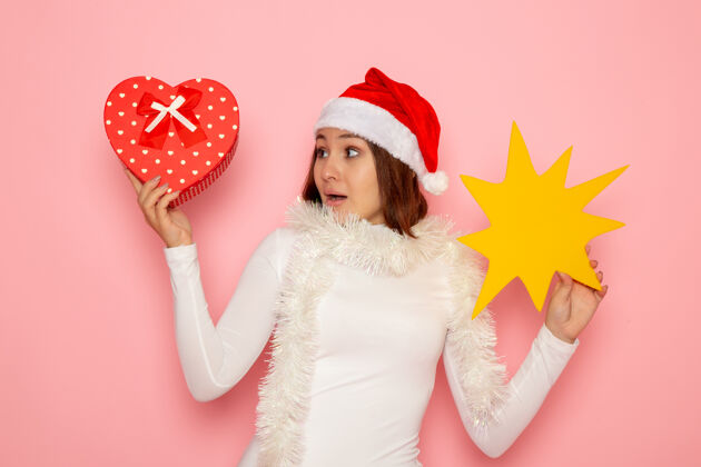 年轻的女性正面图年轻女性手持黄色的身影 呈现在粉色墙壁上雪地圣诞色节日新年时尚圣诞节帽子风景