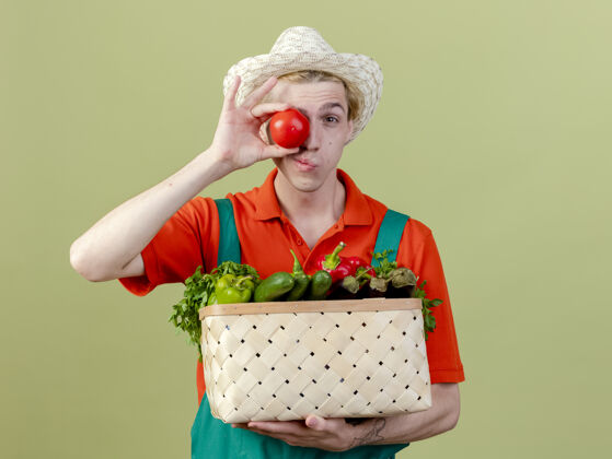 花园年轻的园丁穿着连体衣 戴着帽子 手里拿着装满蔬菜的箱子 站在明亮的背景下微笑着用西红柿遮住眼睛男人帽子西红柿