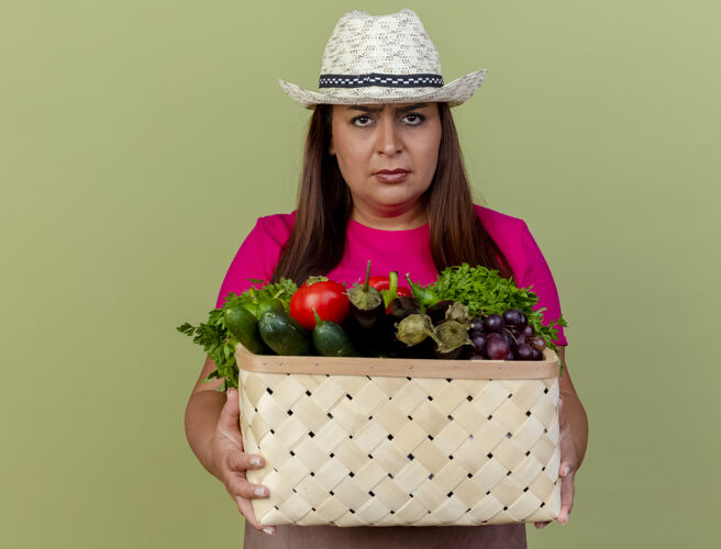 脸中年女园丁围着围裙 戴着帽子 手里拿着装满蔬菜的箱子 站在灯光背景下严肃地看着摄像机帽子满的抱着