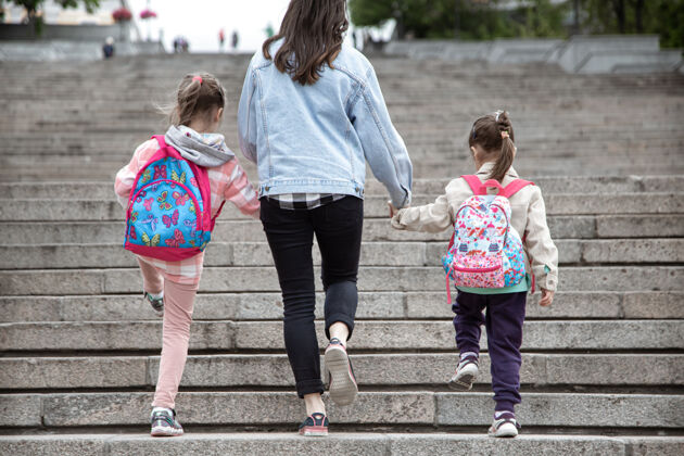 童年家长和小学生手拉手两个女孩的妈妈背着一个背包开始上课秋天的第一天走路从后面走