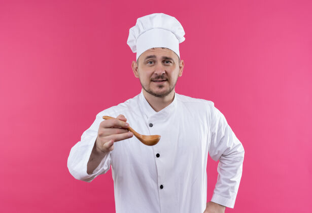 年轻高兴的年轻帅哥厨师穿着厨师制服伸出汤匙孤立在粉红色的空间请粉红色烹饪