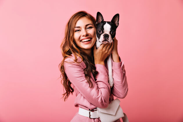 肖像笑容可掬的美女抱着她的小狗姜汁可爱的女孩与法国斗牛犬在粉红色的照片小狗有趣快乐