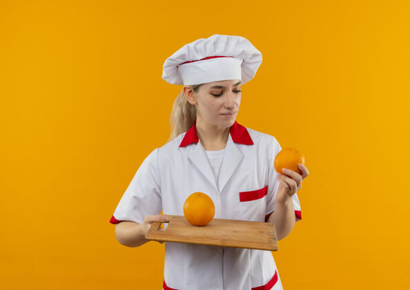 漂亮高兴的年轻漂亮厨师穿着厨师制服拿着橘子和砧板看着橘子把它隔离在橘子的空间里橙子厨师拿着