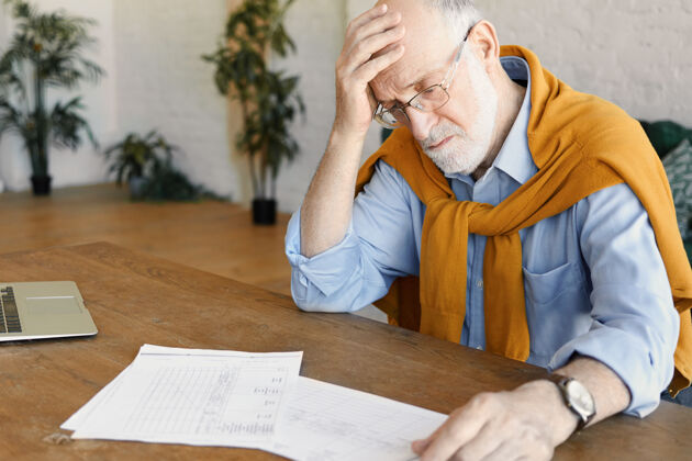 过度工作照片中是一位压力重重 心烦意乱的成熟白人商人 穿着正式的衣服 戴着眼镜 坐在打开的笔记本电脑前 研究文件 面对财务问题 手放在光头上办公桌老人疲惫
