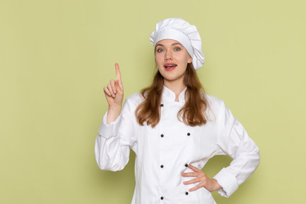 制服身着白色厨师服的女厨师在绿色墙壁上抬起手指的正视图手指正面专业