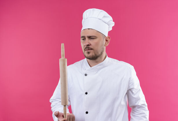 厨师年轻帅气的厨师穿着厨师制服拿着擀面杖看着它孤立的粉色空间年轻制服拿着