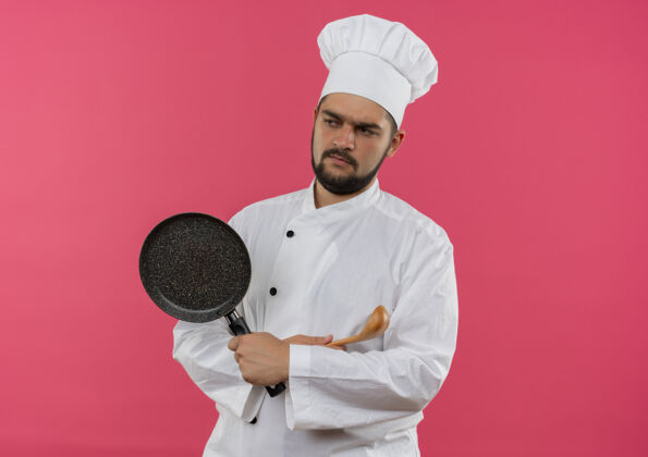 平底锅穿着厨师制服的年轻男厨师站在那里 闭着身子 拿着勺子拿着煎锅 看着粉色空间里孤立的一面侧持有粉红色