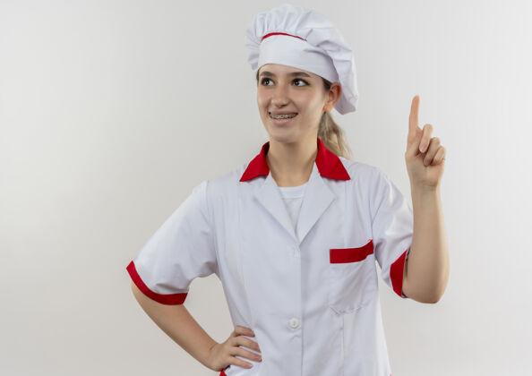 年轻年轻漂亮的厨师 身着厨师制服 戴着牙套 微笑着抬起手指 把手放在腰上 看着被隔离在空白处的一边厨师腰漂亮
