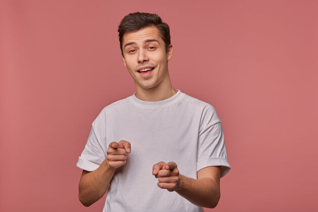 有趣一个年轻开朗的人的肖像 穿着一件空白的t恤 用手指着 站在粉红色的地面上 面带微笑脸快乐年轻