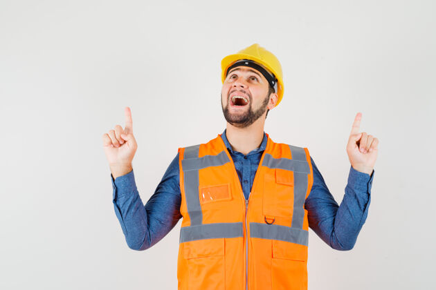 工人年轻的建设者指着手指在衬衫 背心 头盔 看起来很感激 前视图工厂安全帽建筑物