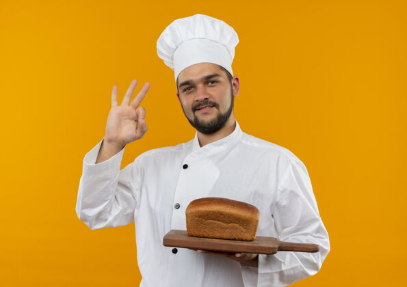 厨师身着厨师制服的年轻男厨师手拿着放着面包的砧板 在橙色的空间里做着“ok”的手势做着高兴好的