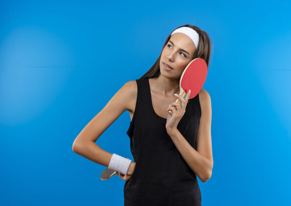 蓝色体贴的年轻漂亮的运动女孩戴着头带和腕带 拿着乒乓球拍 手放在腰上 看着蓝色空间里孤立的一面看起来运动侧面