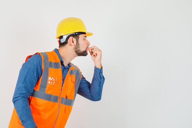 肖像年轻的建筑工人穿着衬衫 背心 头盔 亲吻手指 看起来很高兴工作建筑帽子