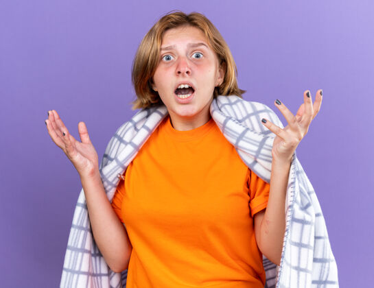 手臂困惑不健康的年轻女子裹着温暖的毯子 感觉不舒服 患流感 发烧 用温度计测量体温 看起来很担心 举起手臂站在紫色的墙上不健康担心痛苦