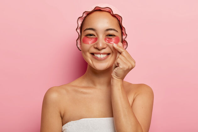 请高兴的韩国女人的近景肖像画做了一个像标志 交叉拇指和食指 送上爱 戴上粉红色的浴帽 在眼睛下面做美容治疗 敷上胶原蛋白贴片保湿赤膊治疗完美