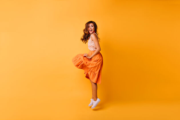 欢呼穿着橙色裙子的浪漫美丽女士的全长肖像在黄色上跳跃的时尚无忧无虑的女孩狂喜女性有趣