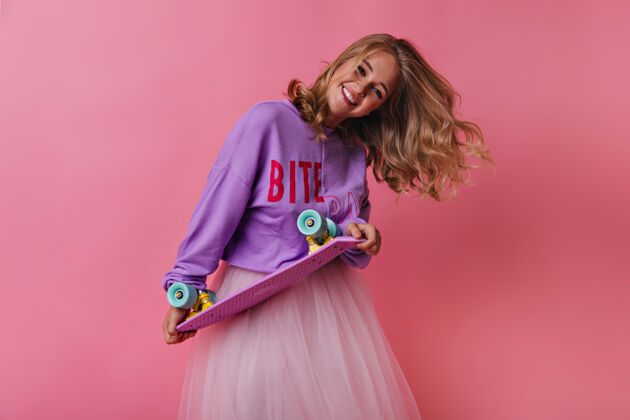 年轻人穿着休闲衬衫的金发女孩拿着滑板笑着迷人的女模特在粉色上表达真诚的情感运动快乐肖像