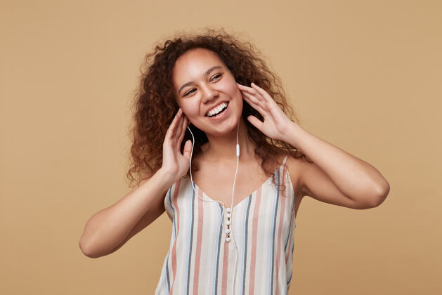 上衣年轻快乐的棕色头发卷曲的女人的肖像 举起双手放在耳机上 愉快地微笑 在米色条纹上衣上摆姿势室内音乐休闲
