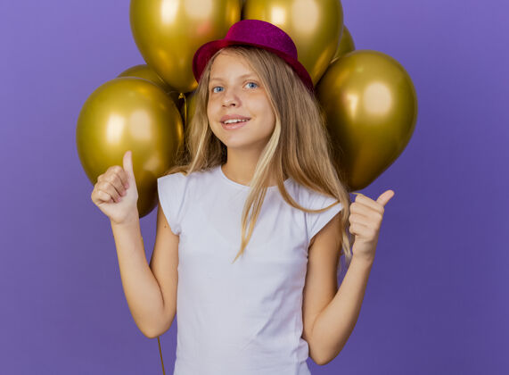 看戴着节日礼帽的漂亮小女孩和一群巴龙看着相机微笑着竖起大拇指 紫色背景下站着生日派对的概念帽子派对紫色