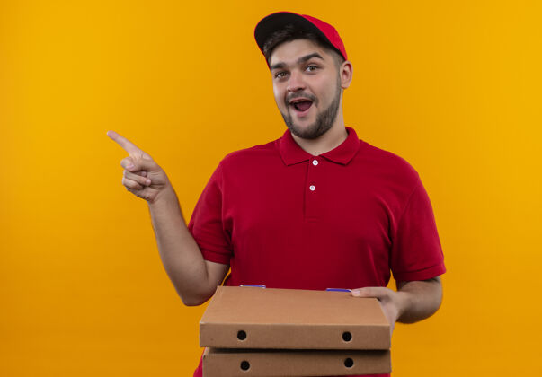 送货年轻的送货员身穿红色制服 戴着帽子 手指指着拿着披萨盒的一边 满面笑容制服站着指着
