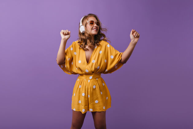 女性可爱的棕褐色女孩 戴着白色耳机 在紫色的舞台上跳舞身着黄色服装的优雅金发女模特在室内欣赏音乐的肖像服饰耳机肖像