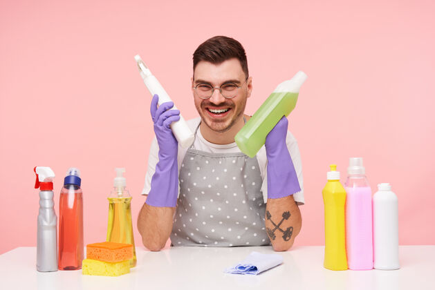 男人室内写真：一个快乐的年轻漂亮的短发黑发男人 手里拿着几瓶家用化学品 坐在粉红色的椅子上开心地笑着抬高清洁室内