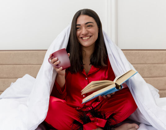 咖啡穿着红色睡衣的年轻美女坐在床上 裹着毛毯 端着一杯咖啡和一本书 在明亮的背景下 愉快地微笑着躺在卧室里坐着欢快快乐