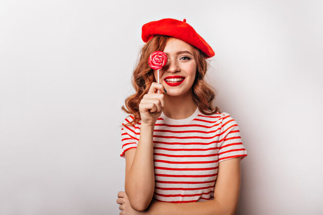 表情穿着红色贝雷帽的温文尔雅的女孩和棒棒糖合影梦幻的法国女模特拿着糖果站在白色的墙上性感漂亮积极