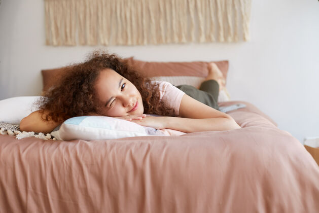 牙齿年轻的黑皮肤可爱的卷发女性肖像躺在床上 看起来很开心 在家里享受阳光和微笑学生美国好