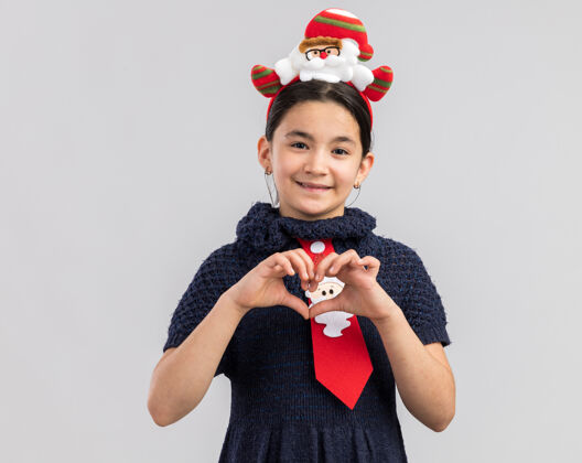 领带小女孩穿着针织连衣裙 头上系着一条红色领带 头上戴着一个有趣的圣诞戒指 面带微笑 用手指做着心形手势新做心