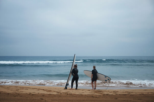 人两个面目全非的冲浪女孩带着她们的长板 一大早就呆在海边看海浪 穿着湿衣 准备冲浪剪影波浪冲浪板