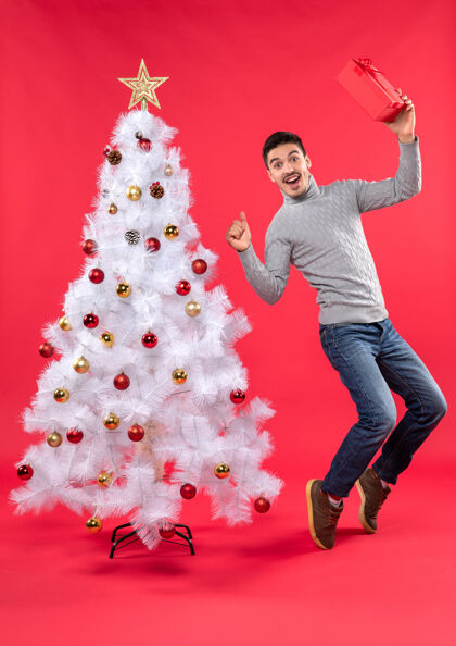 男人俯视图：年轻人站在装饰好的白色新年树旁 手里拿着礼物 在红色的树上跳跃礼物跳舞年轻人