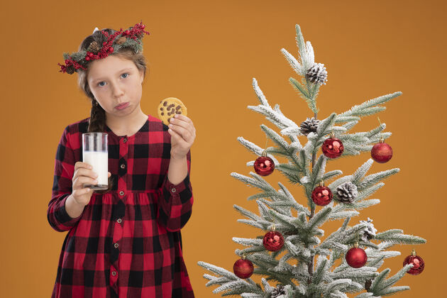 小的悲伤的小女孩戴着圣诞花圈 穿着格子衬衫 手里拿着一杯牛奶和饼干 脸上带着悲伤的表情衬衫悲伤牛奶