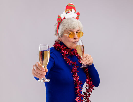 香槟戴着太阳眼镜 脖子上戴着圣诞老人头带和花环的自信的老妇人拿着并喝着几杯香槟新年自信头带