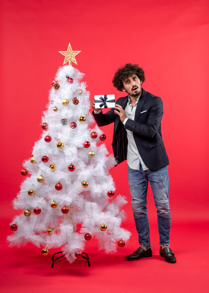 男人惊讶的年轻人拿着他的礼物 站在红色的右侧装饰的白色圣诞树旁沉思圣诞树打扮礼物