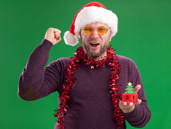 玩具快乐的中年男子戴着圣诞帽 脖子上戴着金丝花环 戴着眼镜 手里拿着圣诞树玩具和日期围着是的看