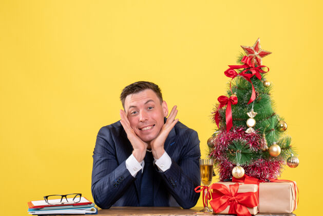 树正面图微笑的男人把手放在下巴上 坐在圣诞树旁的桌子旁 黄色的礼物礼物男人西装