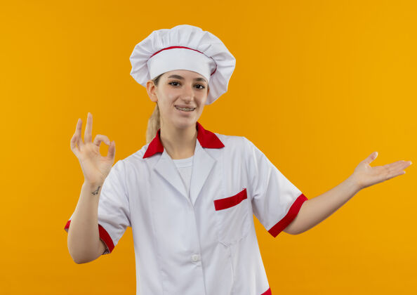 漂亮年轻漂亮的厨师穿着厨师制服 戴着牙套 做着“ok”的手势 空着的手被隔离在橙色的空间里牙科制服烹饪