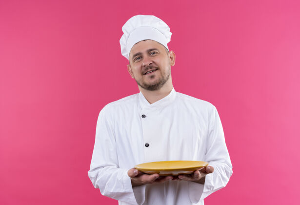 制服高兴的年轻帅哥厨师穿着厨师制服拿着盘子隔离在粉红色的空间里年轻厨师粉红