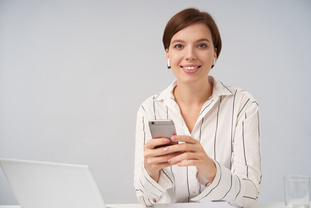 条纹美丽的年轻短发黑发女性 自然妆容 面带微笑 手里拿着手机 穿着条纹衬衫摆出白色的姿势员工姿势智能手机