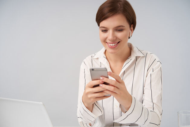 情绪快乐的棕色头发的年轻女性 耳朵里戴着耳机 双手举着智能手机 面带微笑 看着屏幕 摆出白色的姿势电话衣服黑发