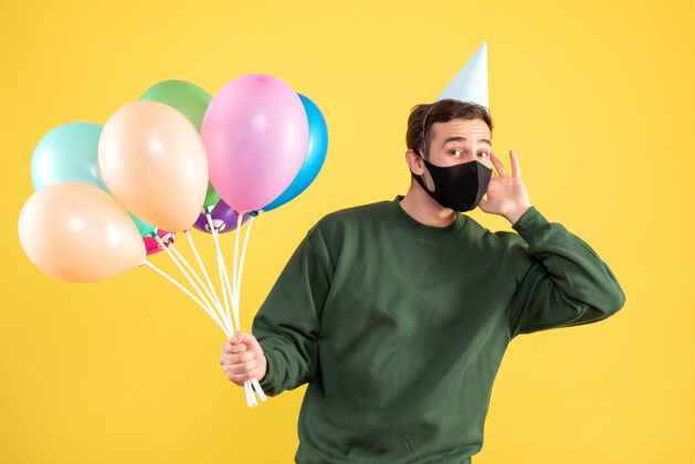 年轻人正面图：戴着黑色面具 站在黄色地面上的彩色气球的年轻人聚会彩色人
