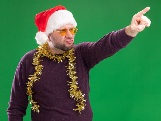 脖子自信的中年男子戴着圣诞帽 脖子上戴着金箔花环 戴着眼镜 看着并指着绿色背景上孤立的一面手指花环中年