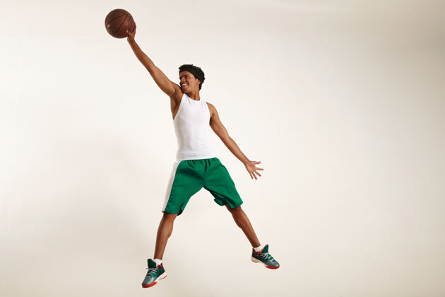 触摸一个穿着白衬衫和绿短裤的快乐的年轻黑人运动员的动作照片跳高抓住一个老式篮球在白色高技术抓