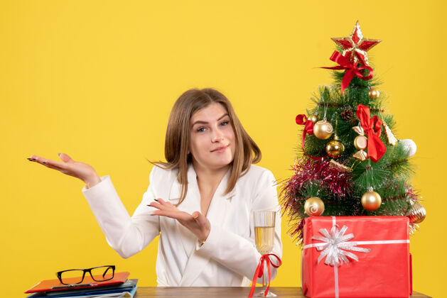 前面正面图：女医生坐在黄色背景的桌子前 放着圣诞树和礼品盒人漂亮女医生