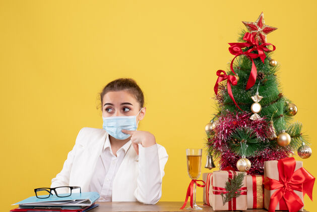 健康正面图女医生戴着黄色背景的无菌口罩坐在圣诞树和礼盒里商务外套病毒