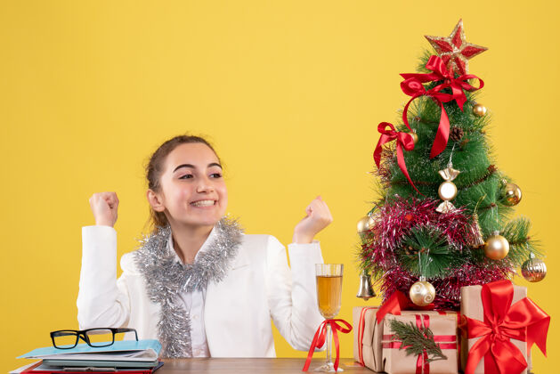 女性前视图：女医生坐在桌子后面 黄色背景上有圣诞礼物和圣诞树护士人圣诞节